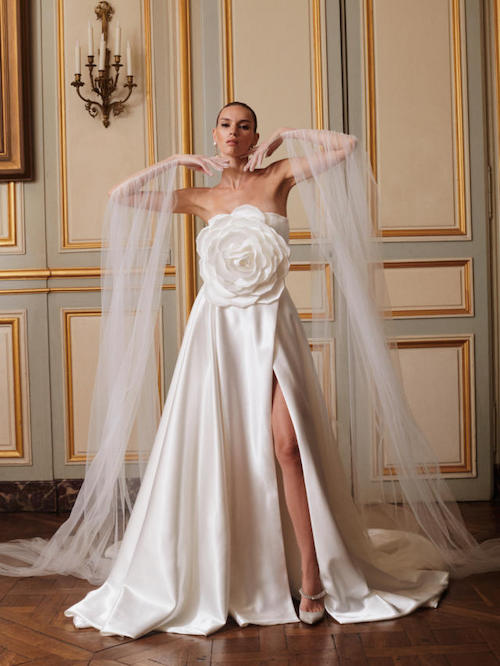 In questa foto una modella in posa con un abito da sposa con spacco 2024 in mikado di seta di Galia Lahv. Splendida la camelia che si irradia sull'intero corpetto del vestito