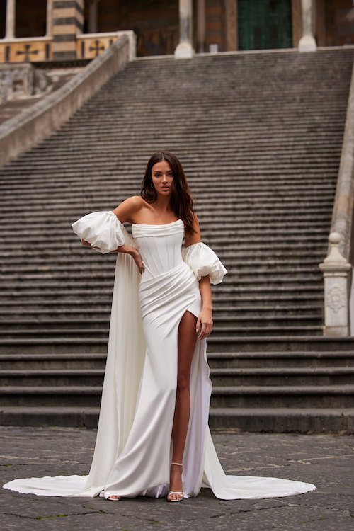 In questa foto una modella indossa un abito da sposa a sirena con spacco di Julie Vino. A completare il look le maniche a palloncino rimovibili, legate sul retro da loro da un lungo strascico