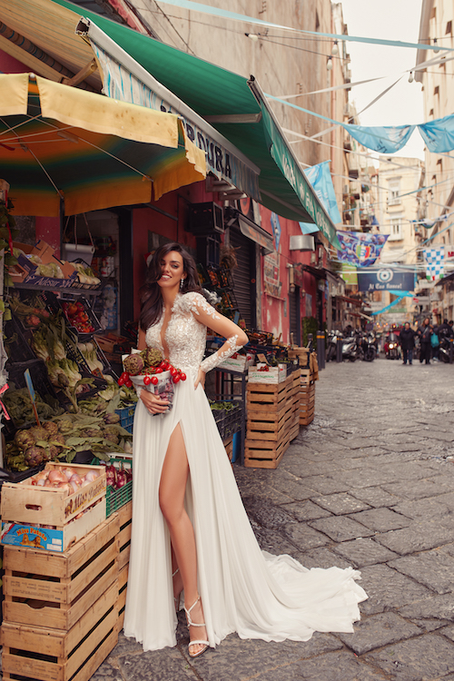 In questa foto una modella posa tra le vie storiche di Napoli, accanto ad una bancarella della frutta, con indossa un vestito da sposa scivolato di Maison Signore: l'abito, che fa parte della collezione Sofia, ha un lungo spacco sulla gamba e il top in pizzo effetto tattoo