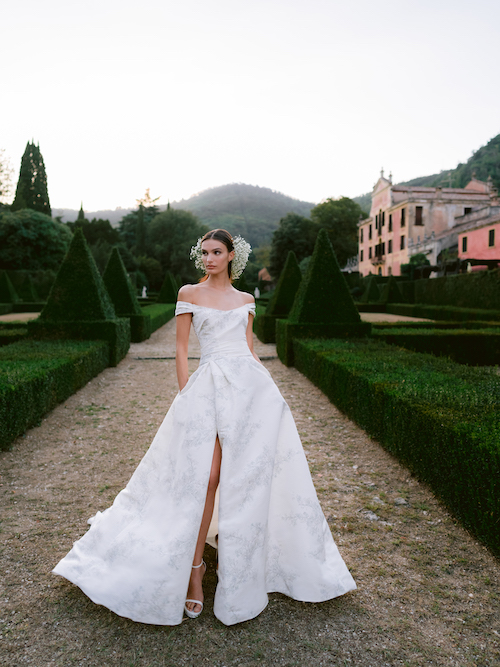 In questa foto una modella posa tra i giardini di una villa italiana con un abito da sposa in mikado di seta con spacco di Monique Lhuillier 2024