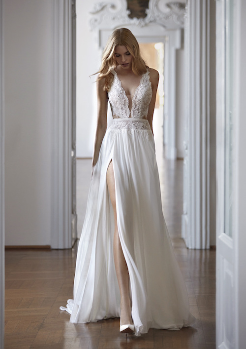 In questa foto una modella attraversa le stanze di un palazzo nobiliare mentre indossa un vestito da sposa scivolato 2024 con spacco, di Nicole Milano