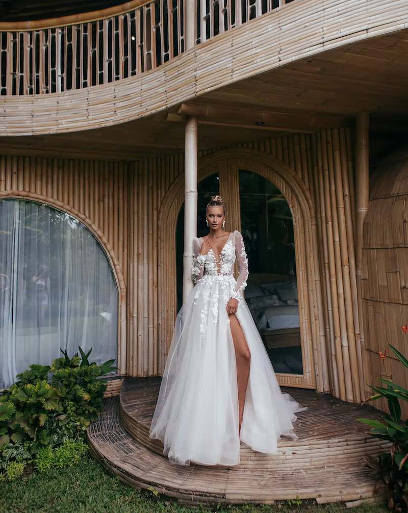 In questa foto una modella posa indossando un abito da sposa Agnieszka Swiatly 2024 con gonna in tulle, spacco frontale, scollo a V e fiori 3D