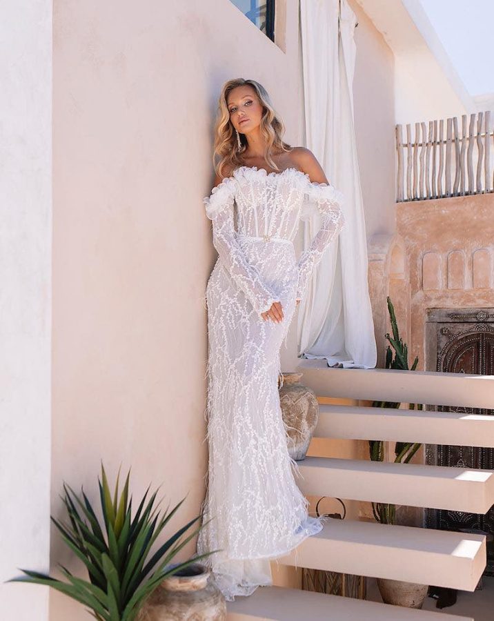 In questa foto una modella indossa un abito da sposa a sirena Agnieszka Swiatly 2024 con ricami di perline e piume, corpetto bustier e maniche lunghe