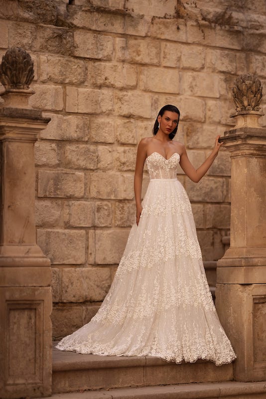 In questa foto una modella posa con un delicato e romantico abito da sposa in pizzo, con bustier e gonna scivolata, di Elena Morar 2024