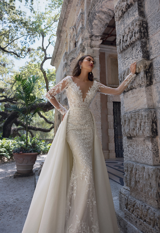 In questa foto una modella posa con un abito da sposa trasformabile di Eva Lendel. Sulla silhouette a sirena, con maniche lunghe in tulle e pizzo, una sovragonna staccabile