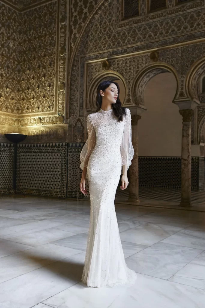 In questa foto una modella posa con un abito da sposa Jenny Packham 2024: bellissime le maniche scivolate balloon in organza trasparente. All over applicazioni lucenti