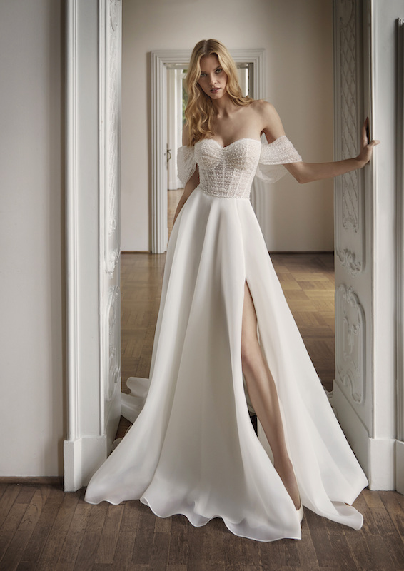 In questa foto una modella indossa un abito da sposa scivolato, con bustier ricoperto da micro perline e maniche corte scivolate, di Nicole Milano. Il vestito, del brand del Gruppo Pronovias, fa parte dei 100 abiti da sposa più belli 2024