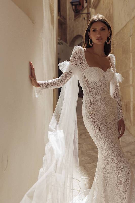 In questa foto una modella indossa un abito da sposa di Pinella Passaro per il 2024: è un vestito a sirena, con maniche lunghe, corpetto steccato e coppe. Il modello è in pizzo all over.