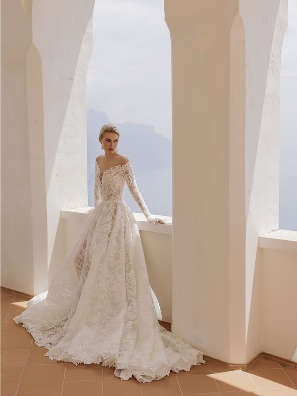 In questa foto una modella indossa un abito da sposa in pizzo all over, con lunghe maniche e scollatura omerale. Il vestito è di Ricca Sposa per il 2024