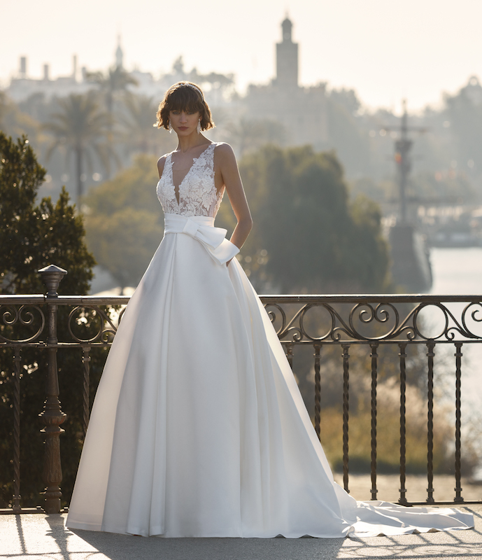 In questa foto una modella posa con un abito da sposa con gonna a trapezio in mikado e top in pizzo di Valerio Luna 2024. Particolare il fiocco che cinge la vita