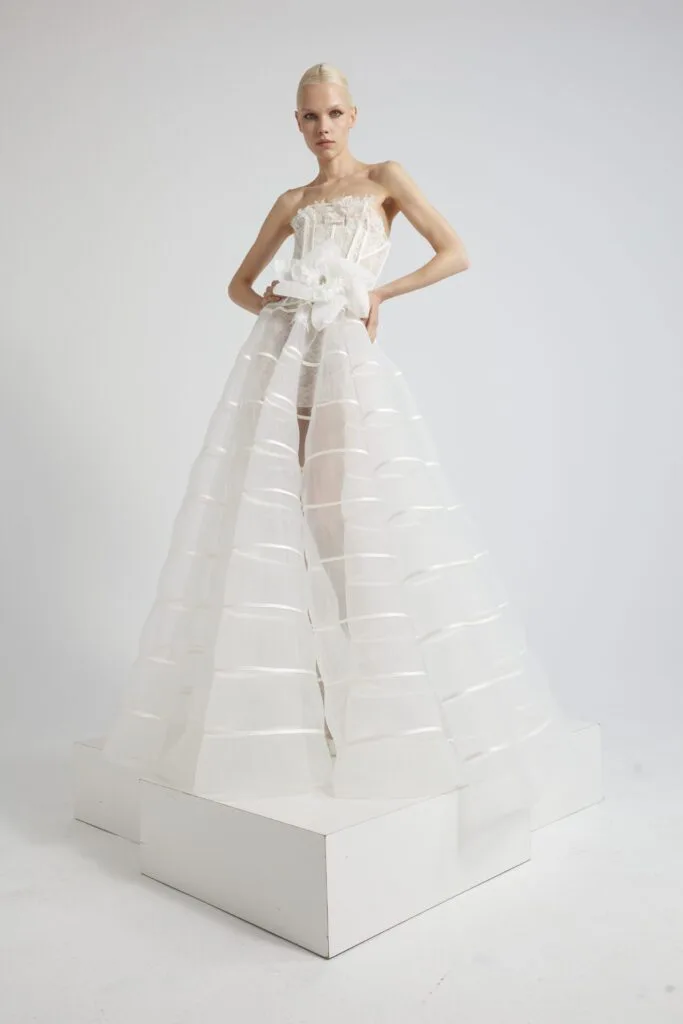 In questa foto una modella indossa un abito da sposa con gonna effetto vedo-non-vedo di YolanCris. Si tratta di una particolare organza a righe orizzontali. Il corpetto è esteccato. In vita un maxi fiocco sempre d'organza bianca