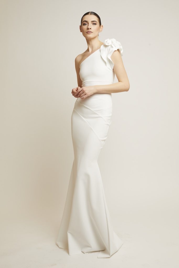 In questa foto una modella indossa un abito da sposa a sirena della collezione di Chiara Boni 2024 in tessuto minimal, monospalla e con fiori 3D sulla spalla sinistra