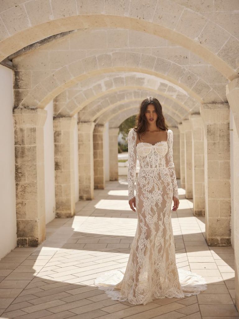 In questa foto una modella indossa uno dei modelli di abiti da sposa a sirena 2024 di Capri Sposa in pizzo e tulle, scollo a cuore e maniche lunghe 