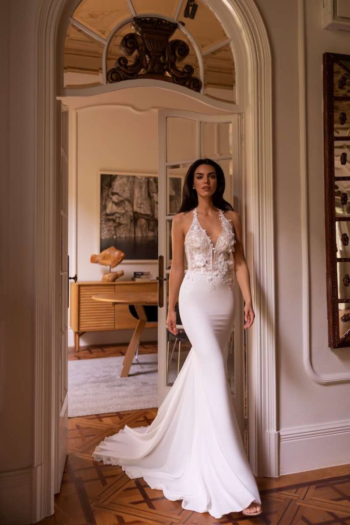 In questa foto una modella indossa uno dei modelli di abiti da sposa a sirena 2024 di Elizabeth Passion con scollo a v, microfiori 3D sul corpetto e gonna avvolgente in crêpe