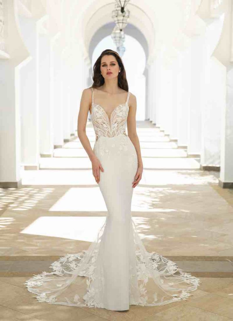 In questa foto una modella indossa uno dei modelli di abiti da sposa a sirena 2024 di Enzoani in crêpe georgette con inserti in pizzo su corpetto e strascico