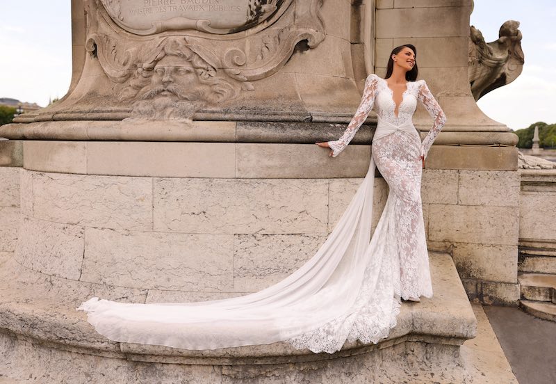 In questa foto una modella indossa uno dei modelli di abiti da sposa a sirena 2024 di Michela Ferriero in pizzo con scollo a V, maniche e lungo strascico