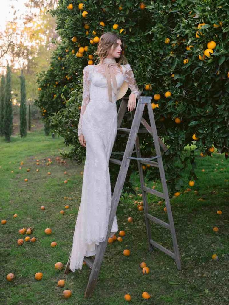 In questa foto una modella indossa uno dei modelli di abiti da sposa a sirena 2024 di Monique Llhuillier con maniche lunghe, collo alto e nastro di velluto di colore cipria al collo