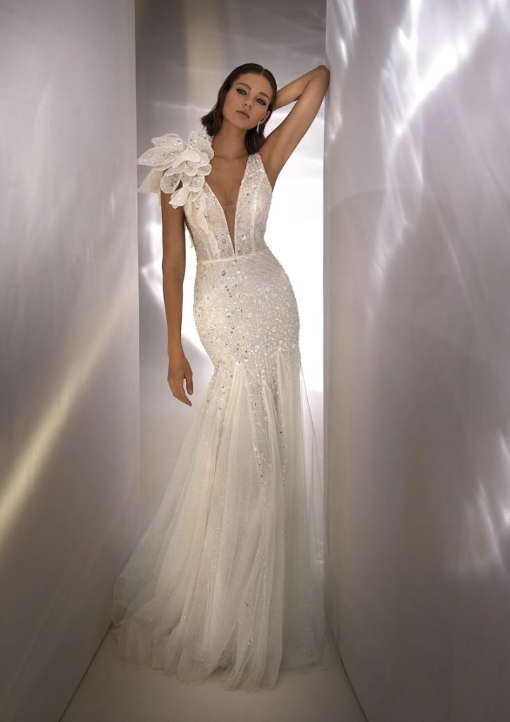 In questa foto una modella indossa uno dei modelli di abiti da sposa a semisirena 2024 di Nicole Milano con dettagli sparkling, scollo a V e maxi fiore sulla spalla destra 