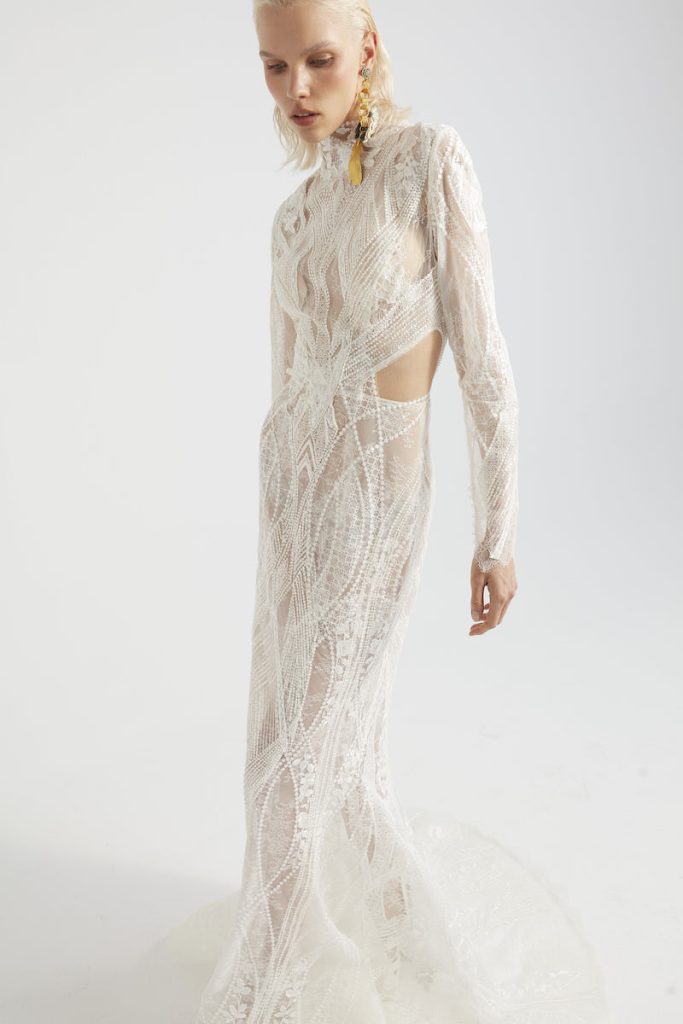 In questa foto una modella indossa un abito da sposa a sirena in pizzo di Yolancris con cut out laterale maniche lunghe