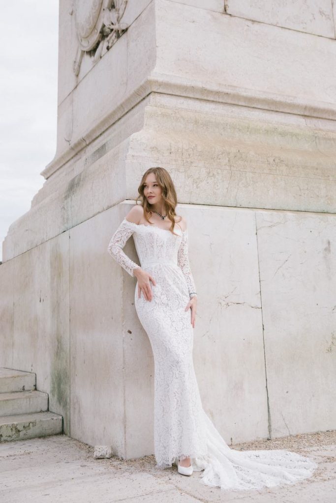 In questa foto una modella indossa un abiti da sposa a sirena Bencivenga 2024 in pizzo con scollo a barchetta e maniche lunghe