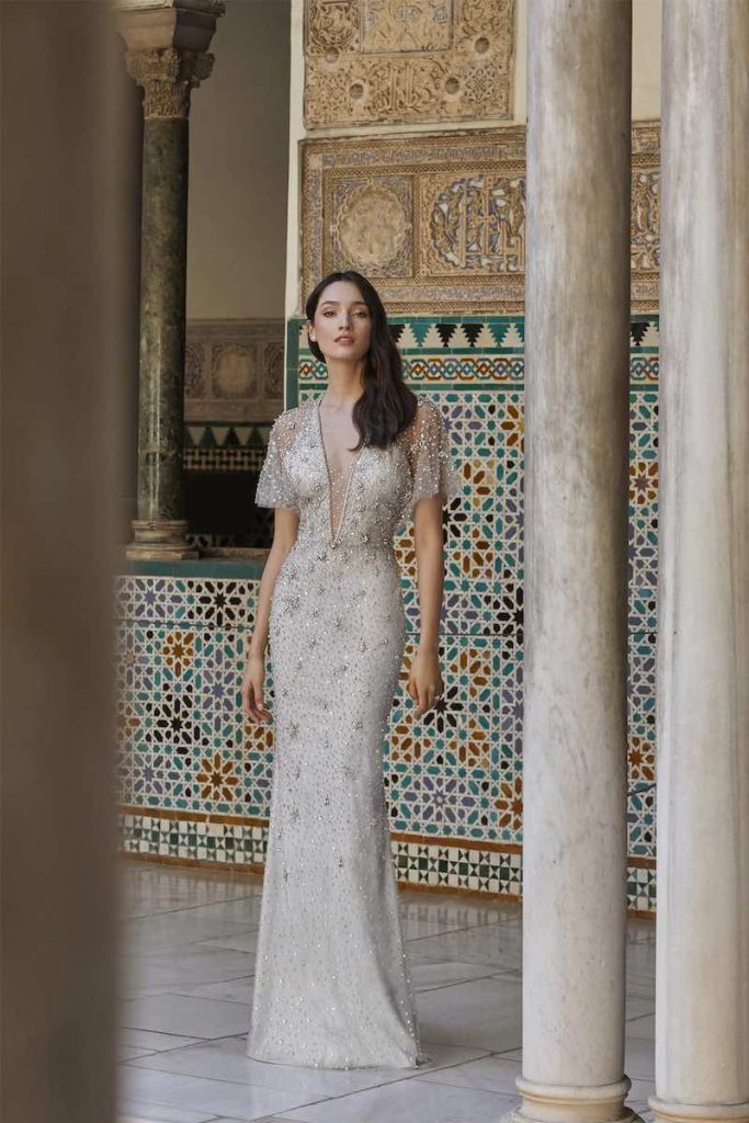In questa foto una modella indossa uno dei modelli di abiti da sposa a sirena 2024 di Jenny Packham con dettagli gioiello, maniche corte e scollo a V