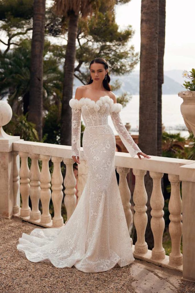 In questa foto una modella indossa uno dei modelli di abiti da sposa a semisirena 2024 di Katy Corso in pizzo e fiori 3D sulla scollatura a barca con maniche lunghe