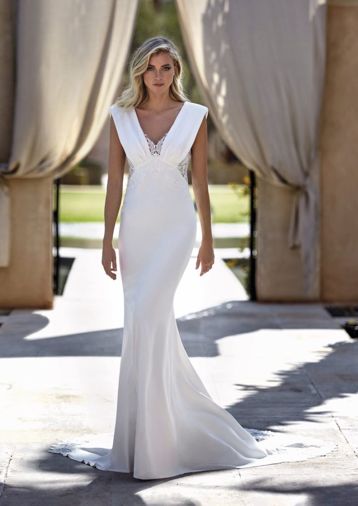 In questa foto una modella indossa uno dei modelli di abiti da sposa a sirena 2024 di Pronovias con scollo a V e inserti a pizzo sul corpetto