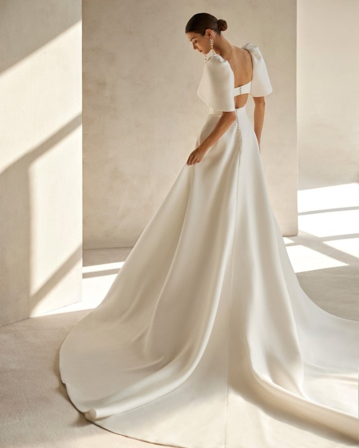 In questa foto una modella inquadrata di spalle indossa un abito da sposa Aire Barcelona 2024 con gonna ampia e strascico in mikado, maniche corte ballon e scollatura sulla schiena