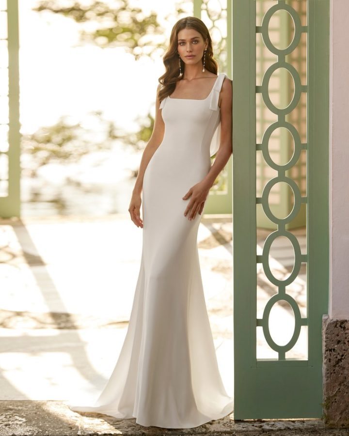 In questa foto una modella indossa uno degli abiti da sposa Aire Barcelona 2024 modello a sirena in crêpe con fiocchi sulle spalle 
