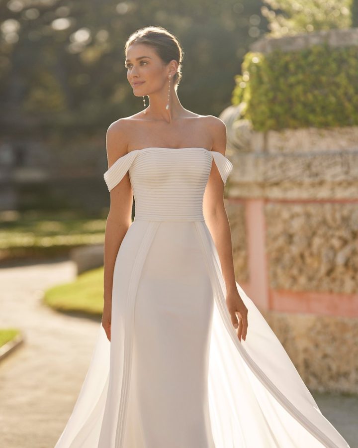 In questa foto una modella inquadrata a tre quarti indossa uno degli abiti da sposa Aire Barcelona 2024 modello a sirena con sopragonna e corpetto a righe 