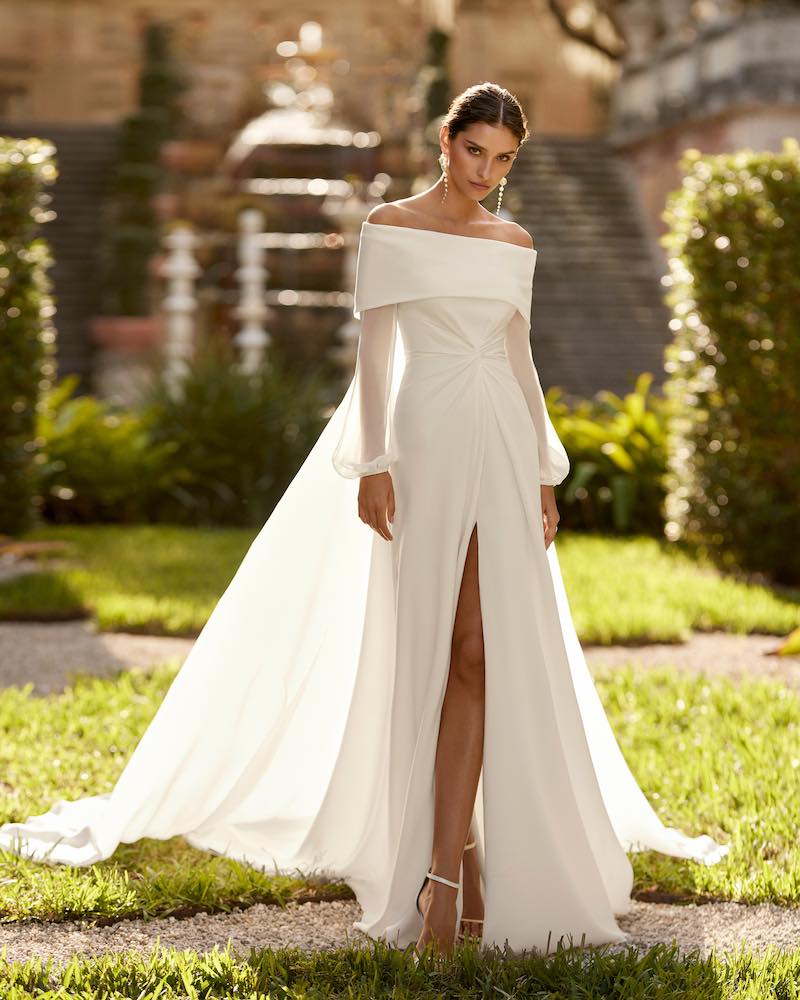 In questa foto una modella indossa uno degli abiti da sposa Aire Barcelona 2024 in crêpe georgette con mantella, spacco frontale, scollo a barca e maniche balloon in chiffon
