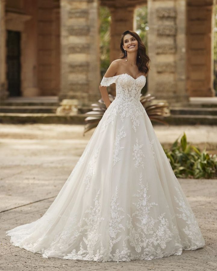 In questa foto una modella indossa un abito da sposa A-line Aire Barcelona Royale 2024 in tulle con ricami in pizzo sull'orlo e ramage sul corpetto e scollatura off-shoulder