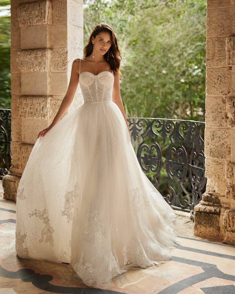 In questa foto una modella indossa un abito da sposa scivolato Aire Barcelona Royale 2024 con gonna in tulle glitterato, corpetto bustier con scollo a cuore e ricami floreali sull'orlo