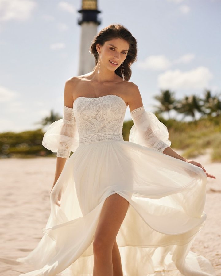 In questa foto una modella inquadrata a tre quarti indossa un abito da sposa Aire Beach Wedding 2024 con spacco frontale, corpetto ricamato  con scollatura dritta e maniche balloon in tulle staccabili