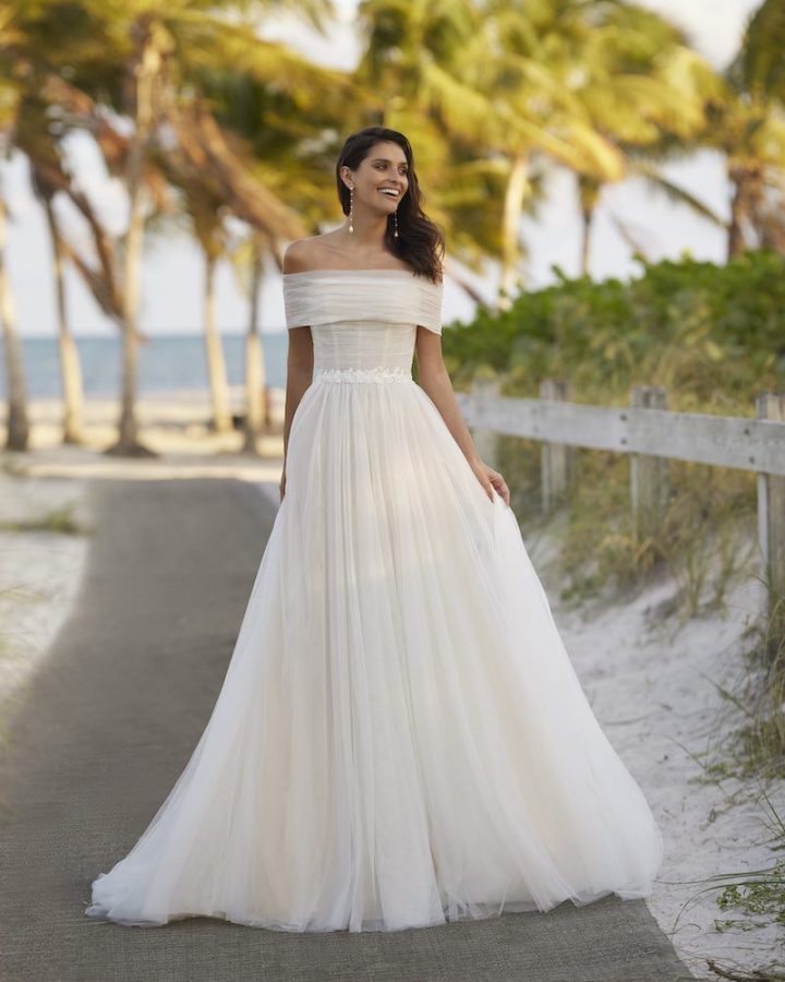 In questa foto una modella posa indossando un abito da sposa Aire Beach Wedding 2024 in tulle con cintura gioiello e bolerino en pendant staccabile