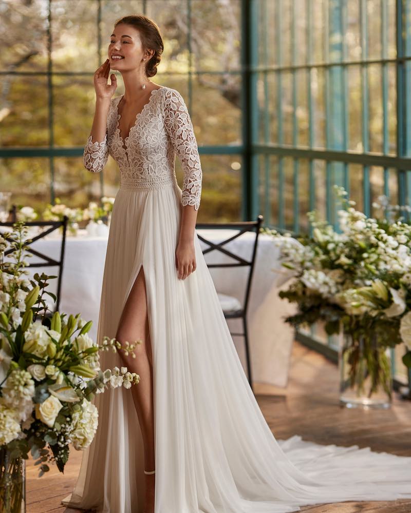 In questa foto una modella posa indossa un abito da sposa scivolato con gonna in chiffon con spacco laterale e corpetto con pizzo floreale con maniche a tre quarti