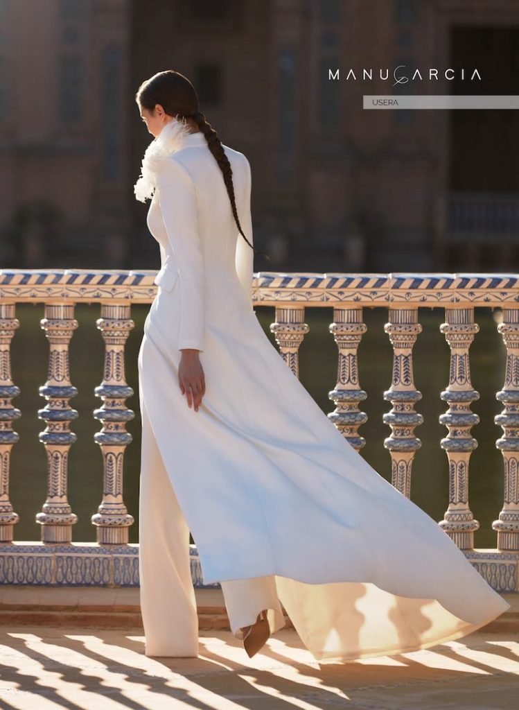 In questa foto una modella di spalle indossa una tuta da sposa Manu Garçia 2024 abbinata ad un cappotto da sposa con piume sul colletto
