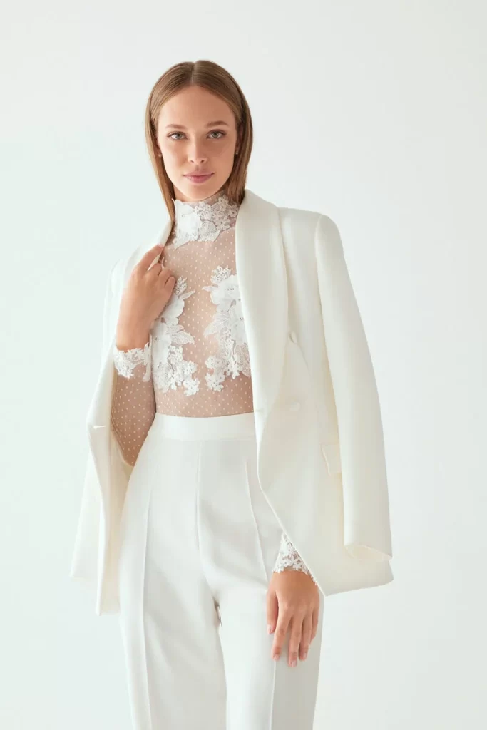In questa una modella indossa uno degli abiti da sposa con pantaloni 2024 di Atelier Emé con corpetto plumetis e ricami di pizzo abbinato ad un blazer en pendant