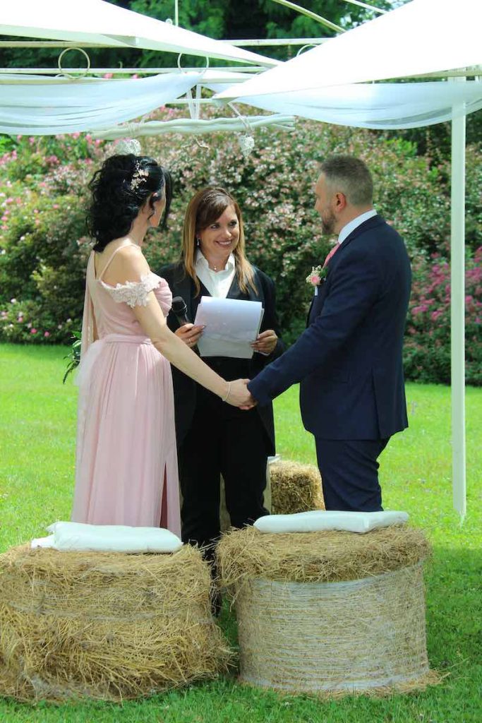 In questa foto due sposi guardano la celebrante Gabriella Maran durante una cerimonia di nozze su un prato. Al posto delle sedie, sono presenti 3 balle di fieno