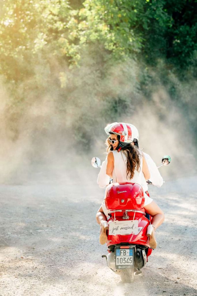 In questa foto di Claudio Fasci due sposi a bordo di una Vespa di colore rosso con il cartello "Just Married" attaccato sul retro corrono su una strada sterrata di campagna. La sposa indossa un casco di colore rosso e, sorridente, è girata verso sinistra 