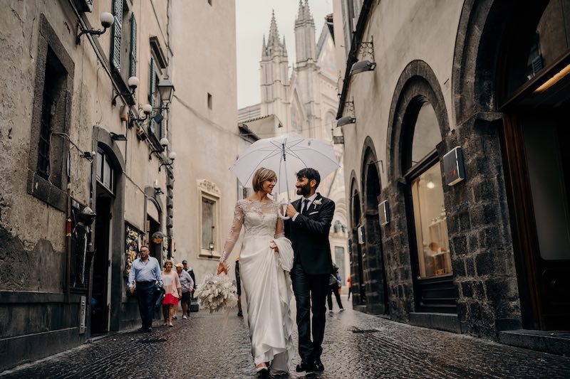 In questa foto due sposi si guardano camminando sotto un ombrello di colore bianco tra le strade di un piccolo paese