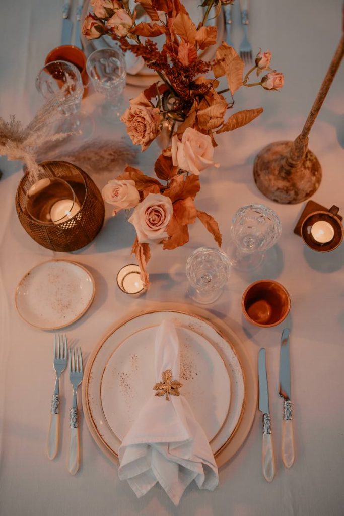 In questa foto scattata dall'alto da Claudio Fasci il dettaglio di una mise en place di matrimonio autunnale decorata con rose e e foglie secche, tovagliato bianco, piatti con bordi colore oro e portacandele in paglia. Il portavogliolo è a forma di foglia di colore oro 