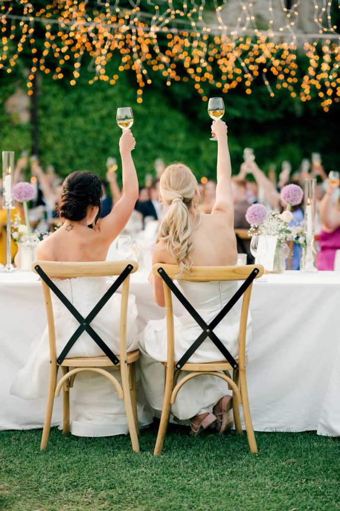In questa foto due spose sedute di spalle alzano due calici di vino per brindare
