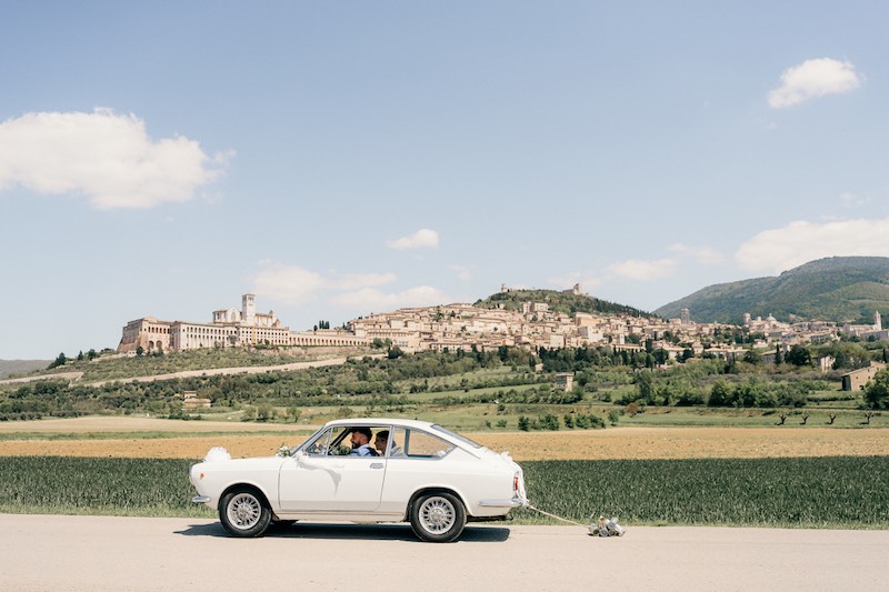 In questa foto due sposi a bordo di un'auto d'epoca di colore bianco sullo sfondo di un paese circondato da campi e prati verdi