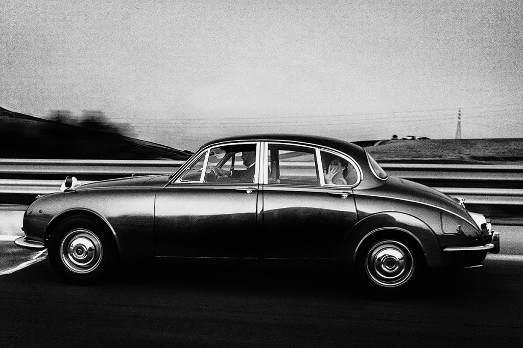 Nella foto in bianco e nero di Antonino Gitto una sposa si dirige all'altare su una macchina d'epoca e saluta dal finestrino 