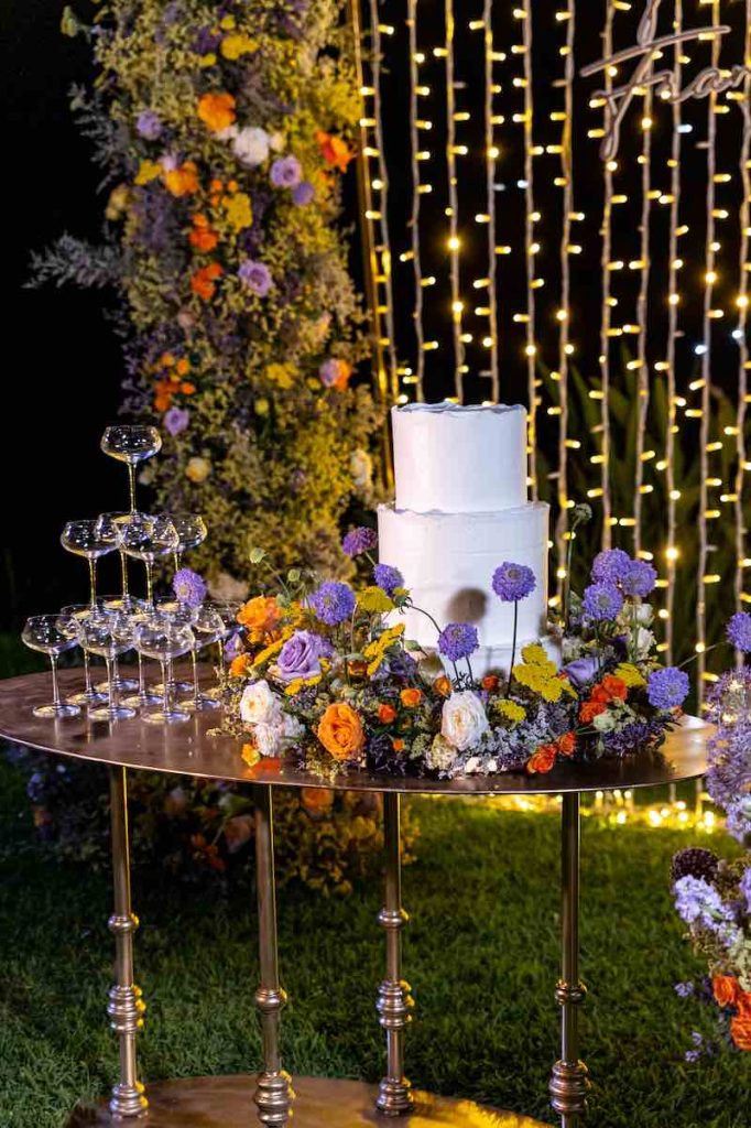 In questa foto il tavolo di una torta nuziale decorato con fiori di colore lilla e arancione e posizionato davanti ad una cascata di luci fredde e un arco di fiori in uno dei matrimoni di Angela De Patto