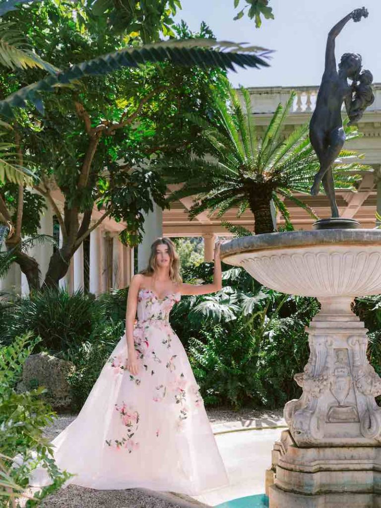 In questa foto una modella posa indossando uno degli abiti da sposa particolari 2024  di Monique Lhuillier con fiori 3D di colore rosa