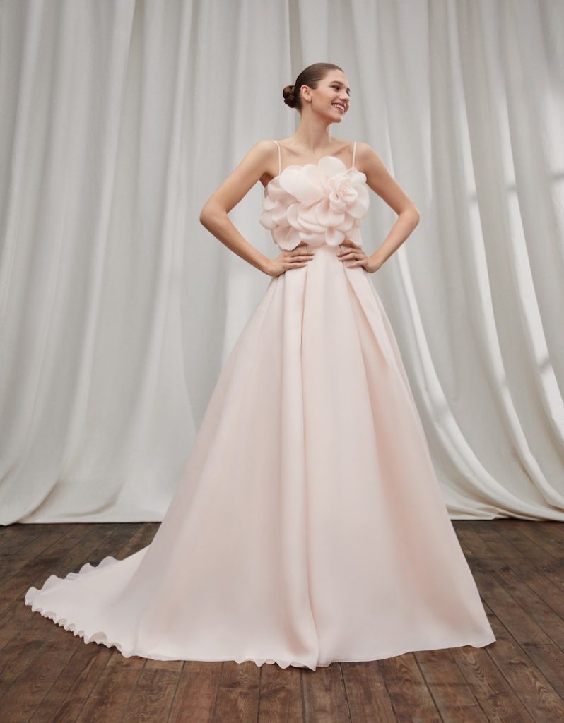 In questa foto una modella posa indossando uno degli abiti da sposa particolari 2024 di Alberto Palatch di colore rosa con maxi camelia sul corpetto