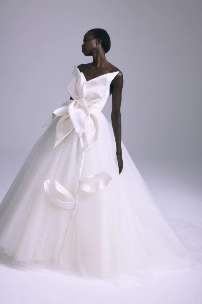 In questa foto una modella posa indossando uno degli abiti da sposa particolari 2024 modello ampio con maxi fiore destrutturato sul corpetto di Amsale