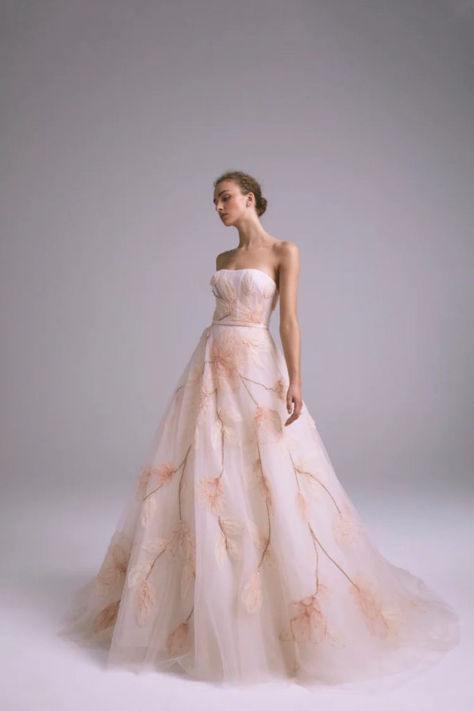 In questa foto una modella posa indossando uno degli abiti da sposa particolari 2024 di Amsale con maxi fiori di colore rosa pesca